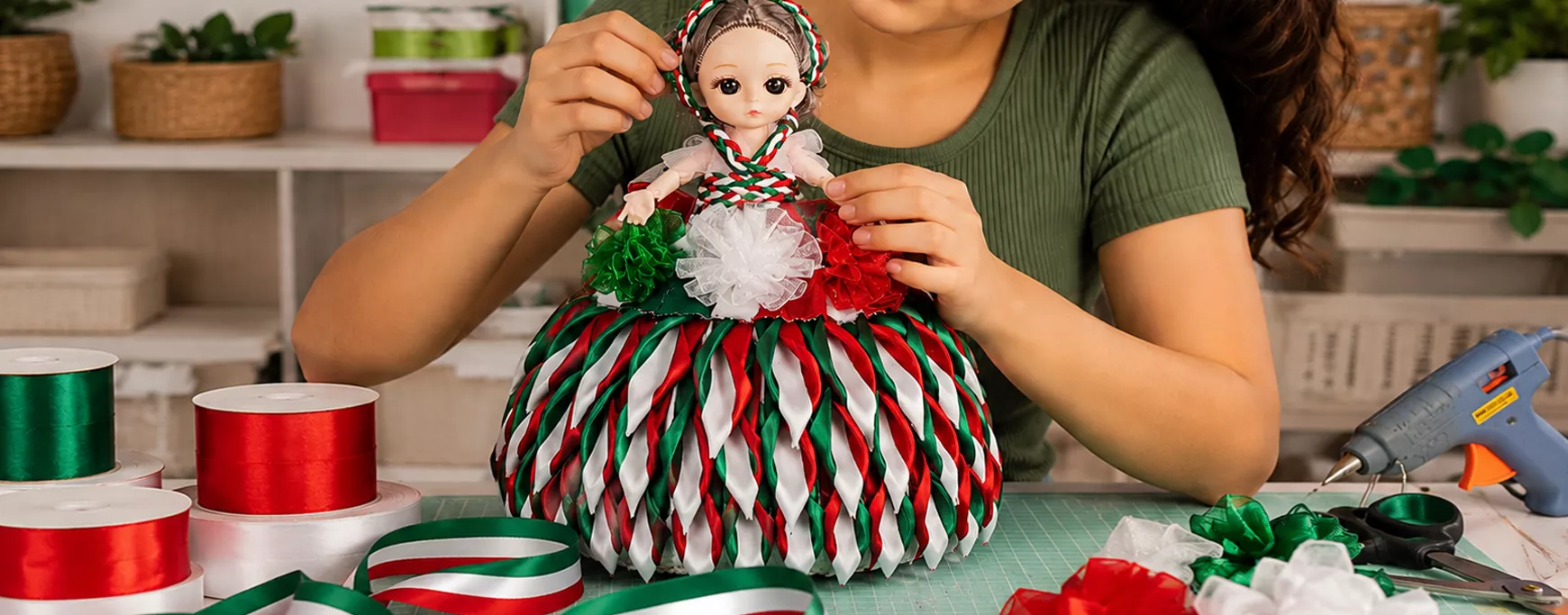 Mujer decorando una muñeca artesanal con listones satinados verdes, blancos y rojos sobre una mesa de trabajo, rodeada de carretes de listón, para las Fiestas Patrias